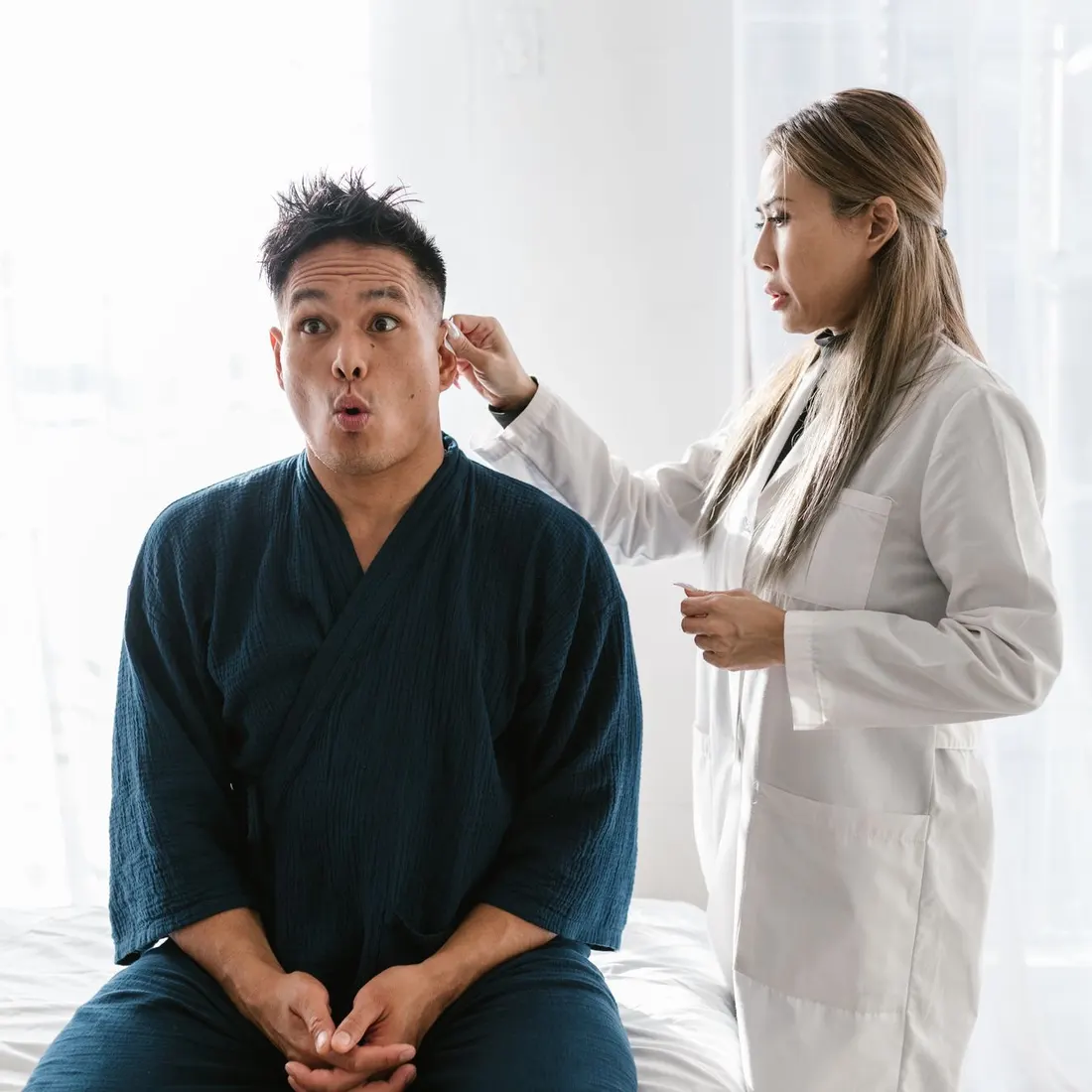 Clinician in a white coat standing beside a seated adult man in a dark blue robe and reaching for his ear, in a bright white room. Photo by RDNE Stock project on Pexels