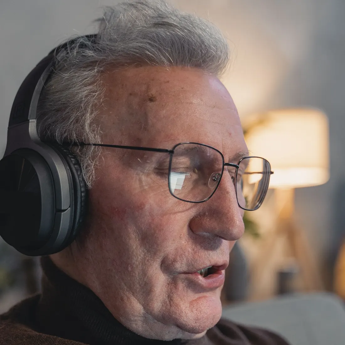Senior man with grey hair and glasses wearing black over-ear headphones in a softly lit room, his mouth open as if speaking. Photo by Tima Miroshnichenko on Pexels