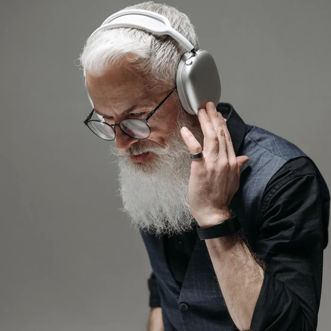 Senior man with a white beard and round glasses holding silver over-ear headphones to his ear, in a dark shirt against a grey background. Photo by Pavel Danilyuk on Pexels