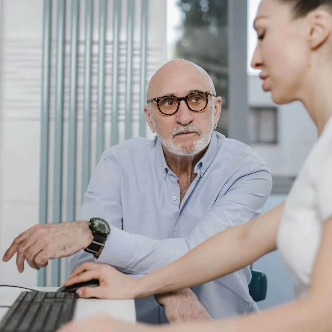 Bald senior man with glasses in a light blue shirt sits at a desk while a woman in a white T-shirt uses a computer mouse beside him. Photo by SHVETS production on Pexels