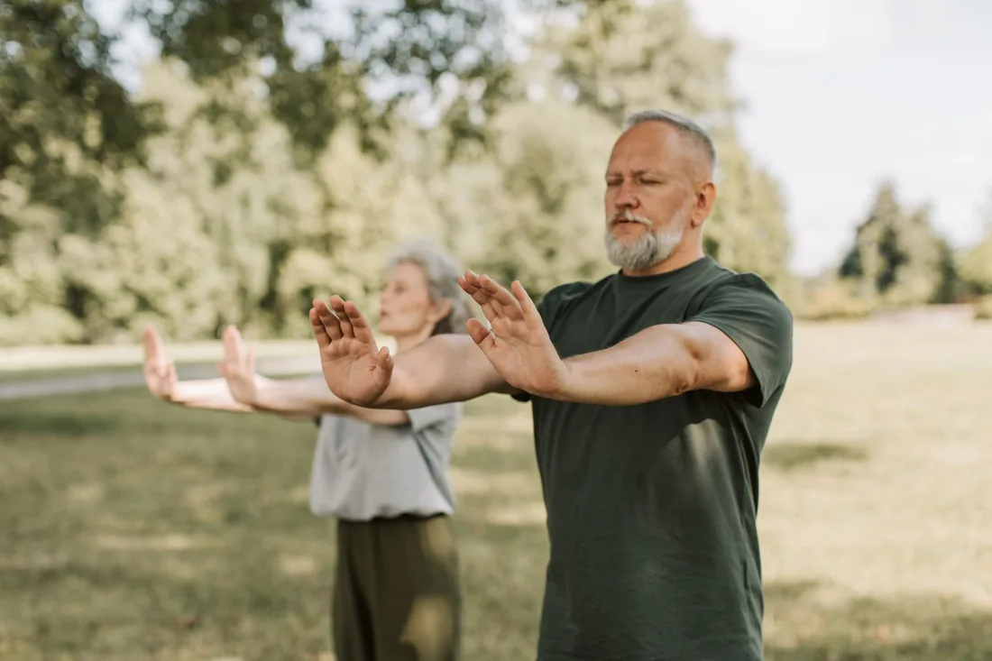 Older man and woman practising tai chi arm movements outdoors in a park. Photo by Vlada Karpovich on Pexels