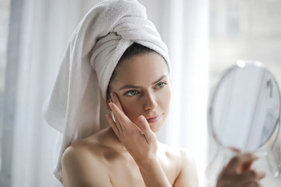 Woman with a towel wrapped around her hair checking her clear skin in a hand mirror. Photo by Andrea Piacquadio on Pexels
