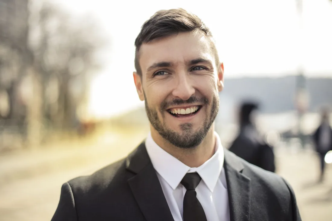 Smiling man in a dark suit with a full head of neatly styled hair outdoors. Photo by Andrea Piacquadio on Pexels