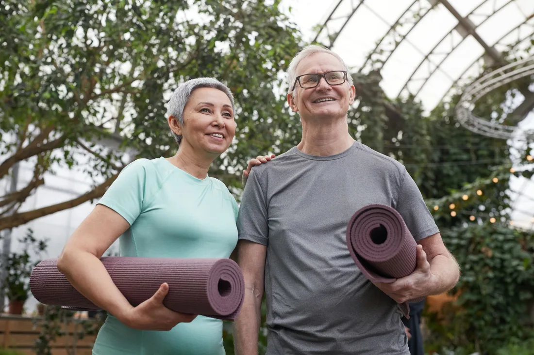 Smiling senior couple carrying yoga mats in a bright garden setting. Photo by Marcus Aurelius on Pexels