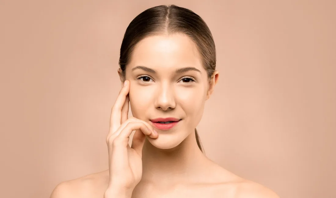 Woman with smooth, glowing skin touching her cheek against a soft peach background. Photo by Shiny Diamond on Pexels