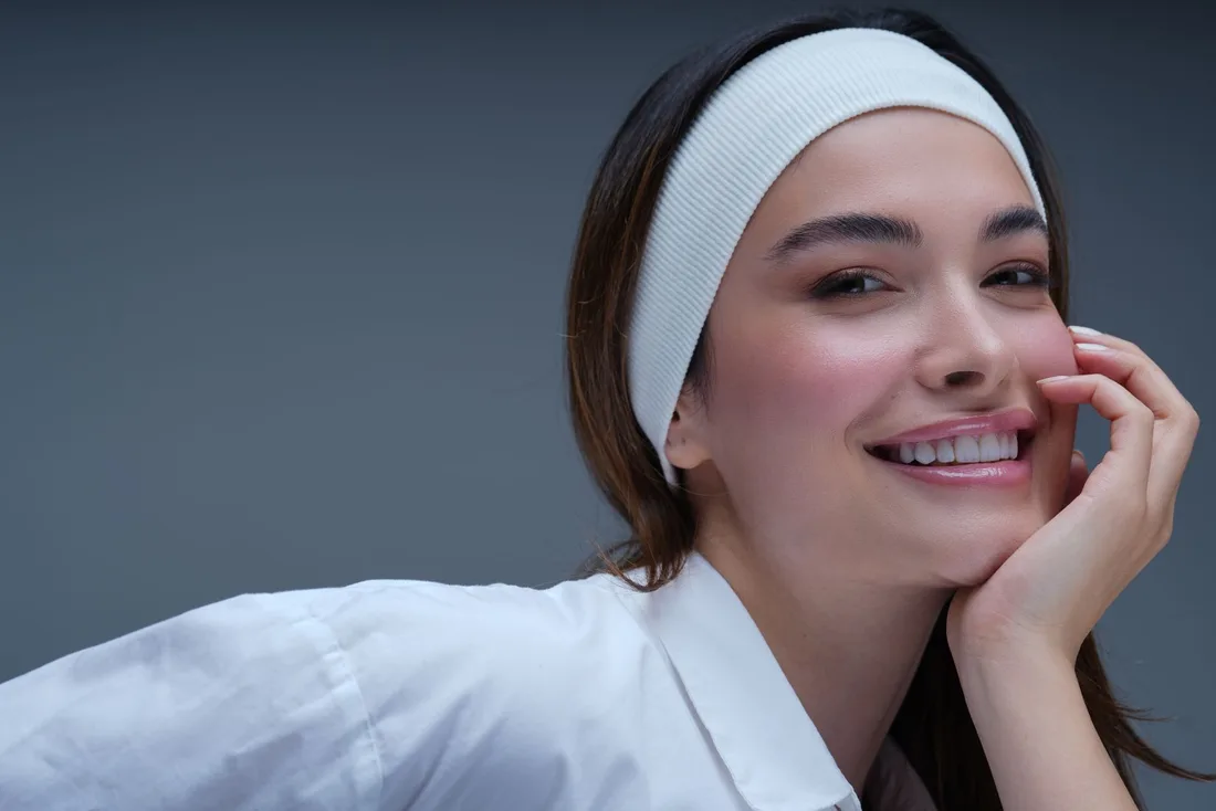 Smiling woman in a white headband with radiant skin resting her chin on her hand. Photo by Farhad Irani on Pexels