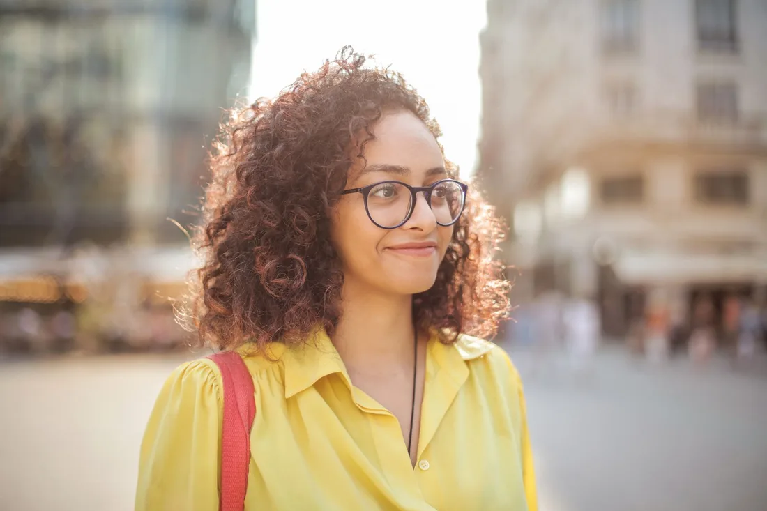 Smiling woman with curly hair wearing round glasses and a yellow top outdoors. Photo by Andrea Piacquadio on Pexels