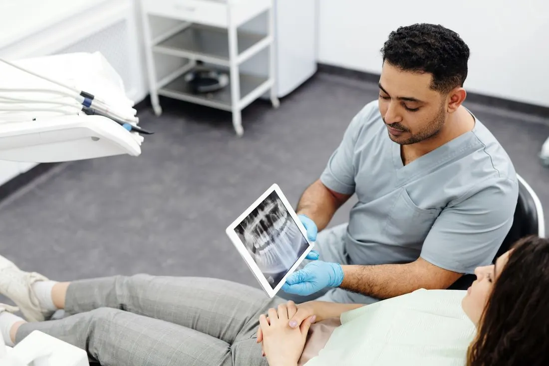 Clinician in grey scrubs and blue gloves holding a tablet that shows a panoramic dental X-ray for a woman reclining in a treatment chair with her hands folded. Photo by Cedric Fauntleroy on Pexels