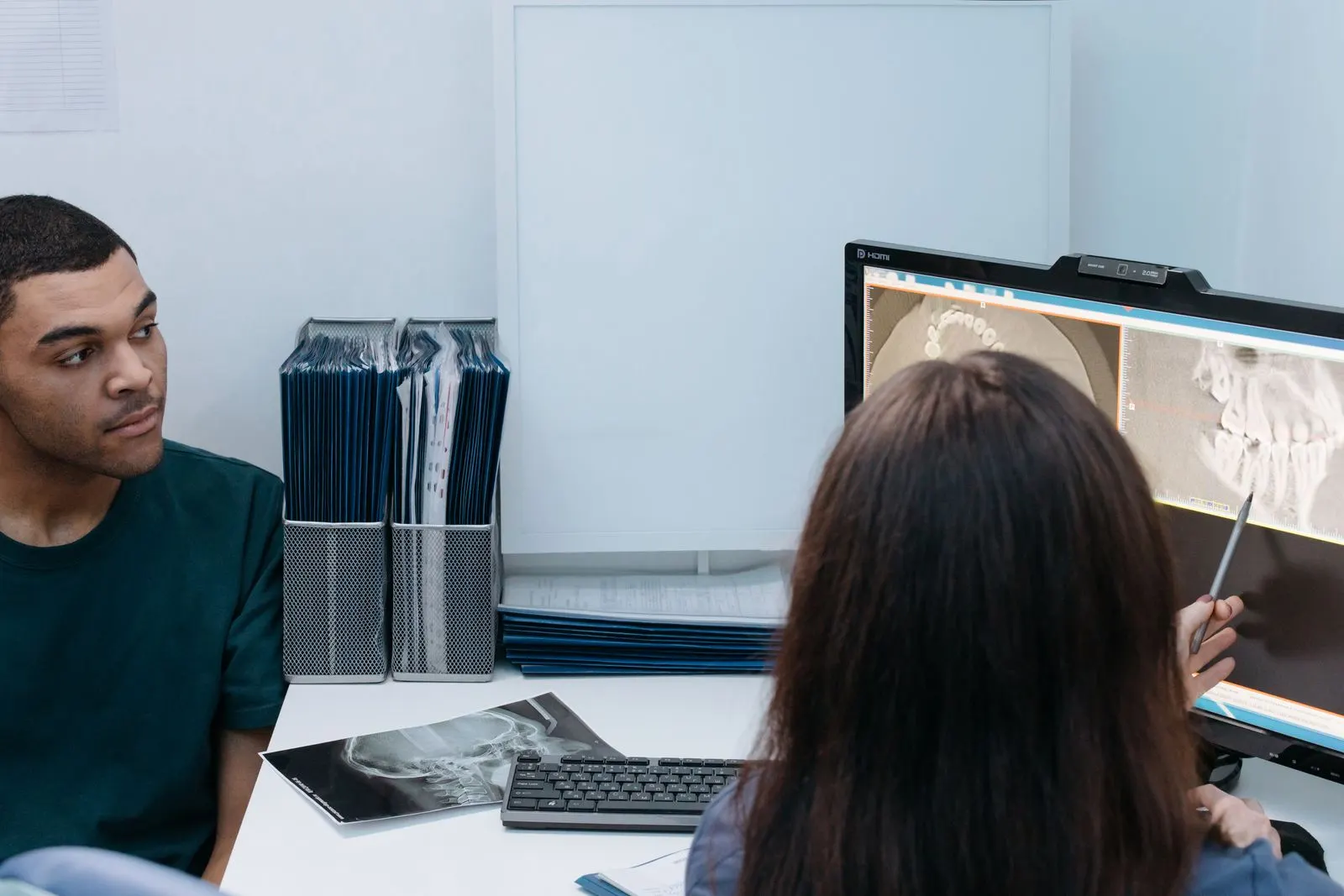 Clinician with long dark hair pointing a stylus at jaw scan images on a desktop monitor while an adult man in a green T-shirt sits across the desk, with a printed skull radiograph and a keyboard between them. Photo by cottonbro studio on Pexels