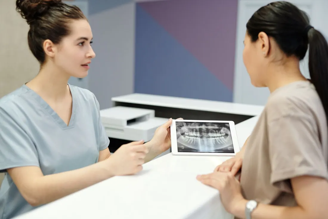 Clinician in light blue scrubs showing a panoramic dental X-ray on a white tablet to an adult woman seated across a white counter. Photo by Cedric Fauntleroy on Pexels