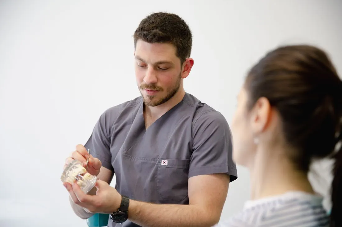 Bearded clinician in grey scrubs holding a clear dental jaw model and pointing at it with a probe while a woman with her hair tied back looks on from the right. Photo by Esma Karagoz on Pexels