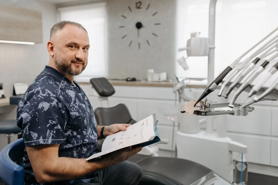 Smiling clinician with a short beard in patterned blue scrubs sitting beside a treatment chair holding an open printed document, with a wall clock and handpieces behind him. Photo by Pavel Danilyuk on Pexels