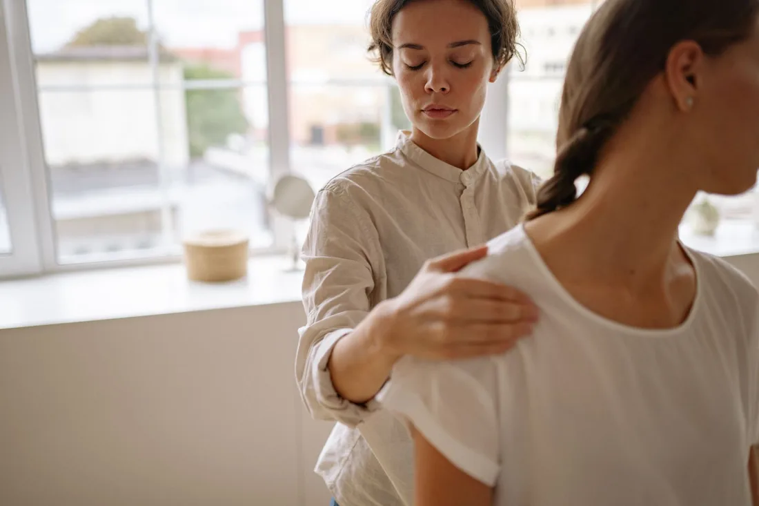 Physiotherapist assisting a seated patient with shoulder movement beside a window. Photo by Yan Krukau on Pexels