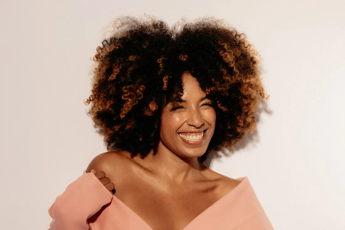 Smiling woman with voluminous curly hair in a bright, warm-toned studio portrait. Photo by Gabriela Brasiliano on Pexels