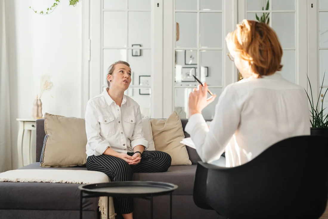 Two women talking in a bright, calm therapy room with a sofa and armchair. Photo by https://kaboompics.com/ on Pexels