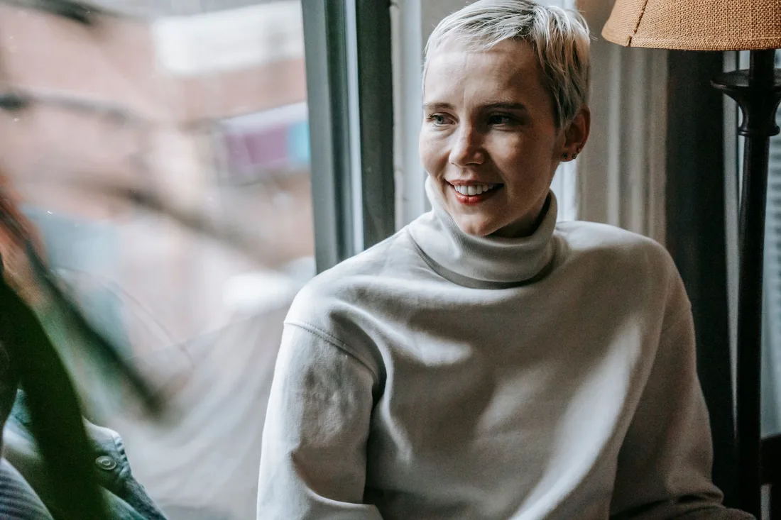 Smiling woman with short grey hair in a cozy sweater looking relaxed by a window. Photo by Gary  Barnes on Pexels