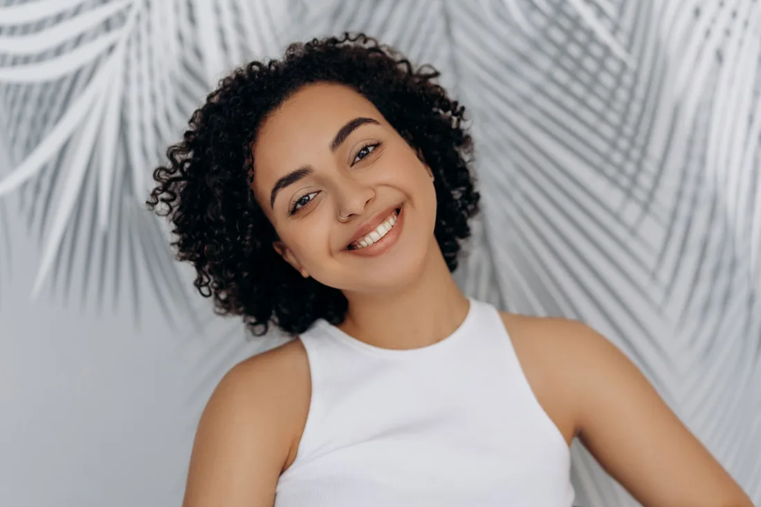 Smiling woman with curly dark hair and clear skin in a bright white top. Photo by Mikhail Nilov on Pexels