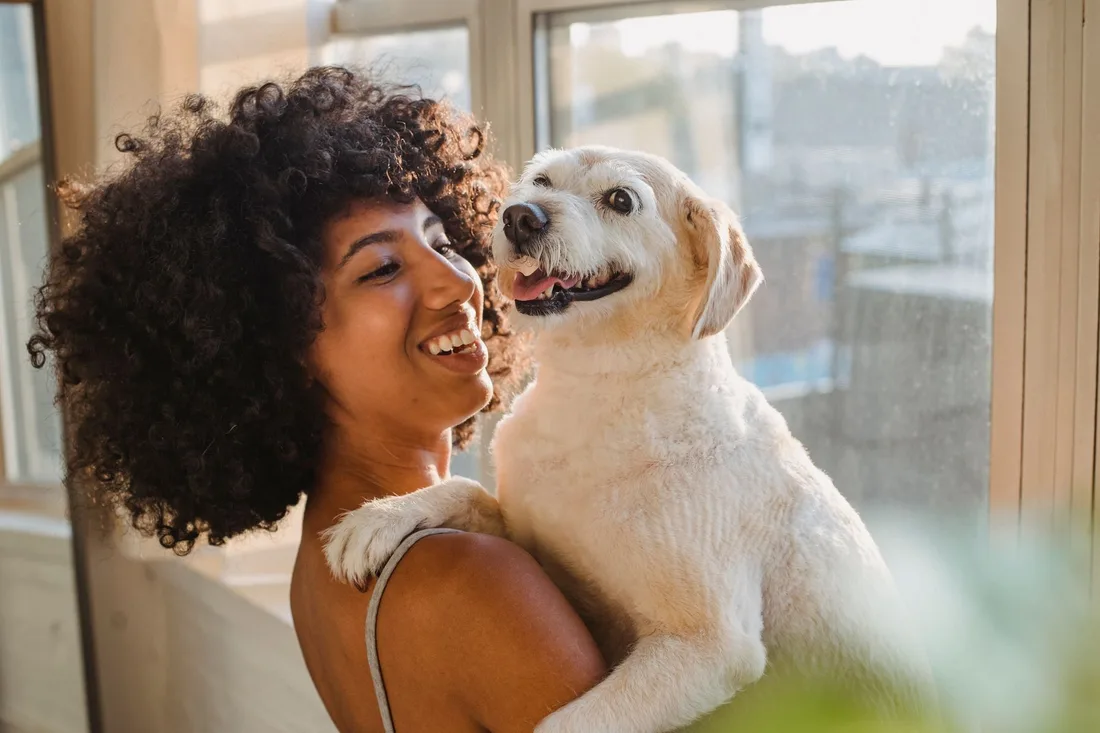 Smiling woman with curly hair lovingly holding her happy dog in a bright room. Photo by Samson Katt on Pexels