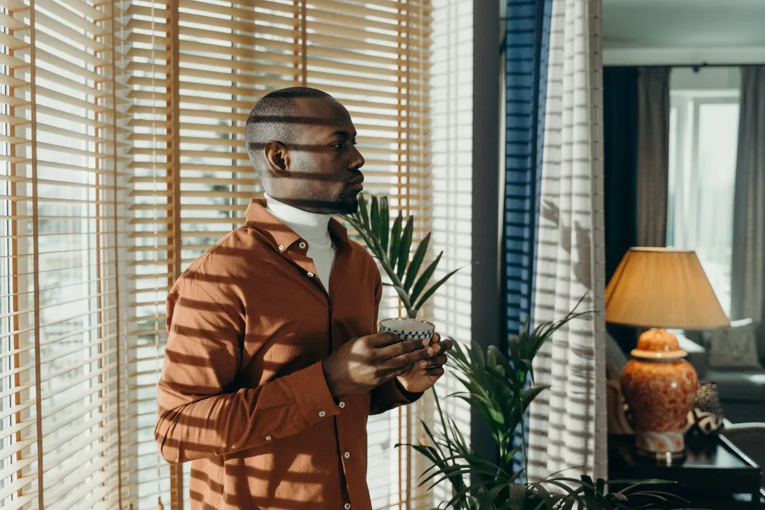 Man in brown shirt holding cup in sunlit room with blinds and plants. Photo by cottonbro studio on Pexels