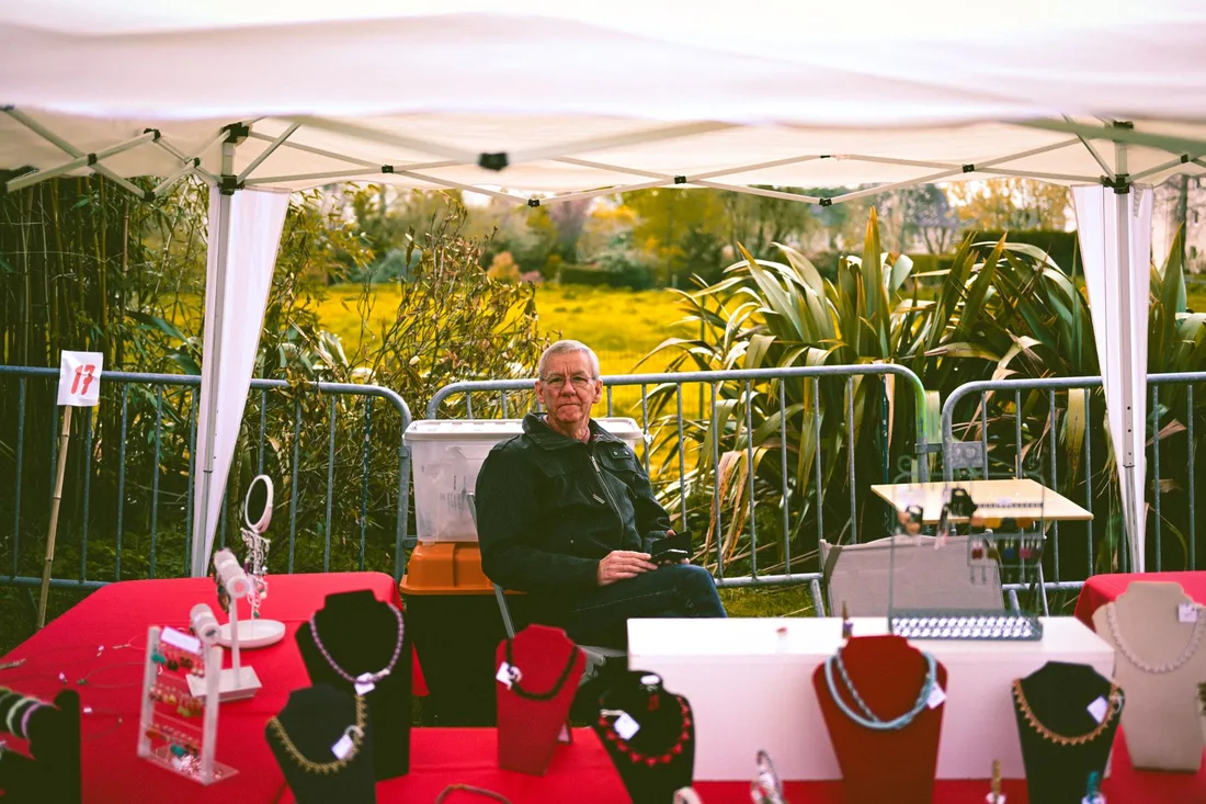 A mature man sits under a canopy in a jewelry booth displaying necklaces outdoors. Photo by Kwami  Heude Izawaki on Pexels