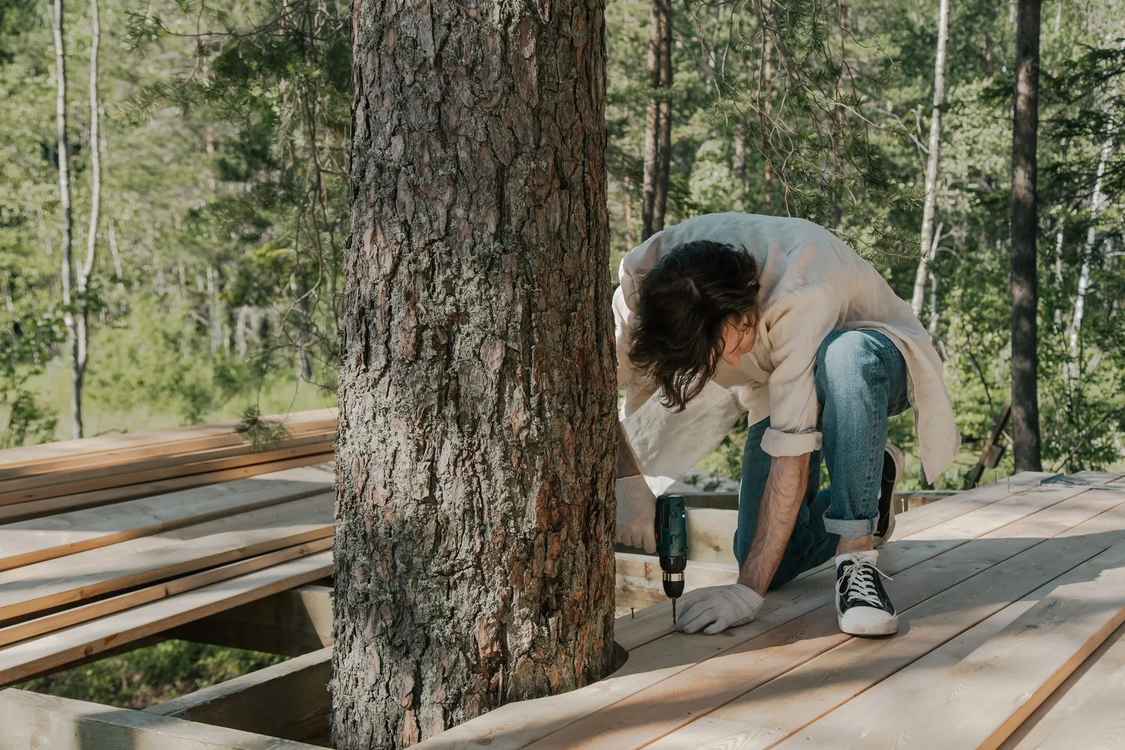 A man performs manual labor using a drill on a forest boardwalk construction project. Photo by Ron Lach on Pexels