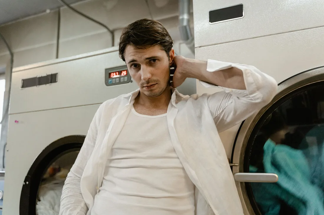 Young man in a white shirt leaning on a washing machine in a laundry facility. Photo by Tima Miroshnichenko on Pexels
