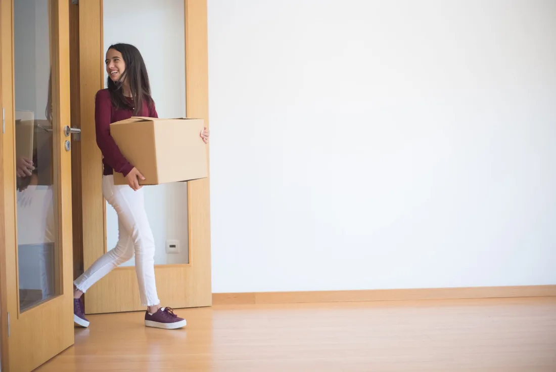 Woman carrying a box into a bright empty room with a new wood floor. Photo by Kampus Production on Pexels