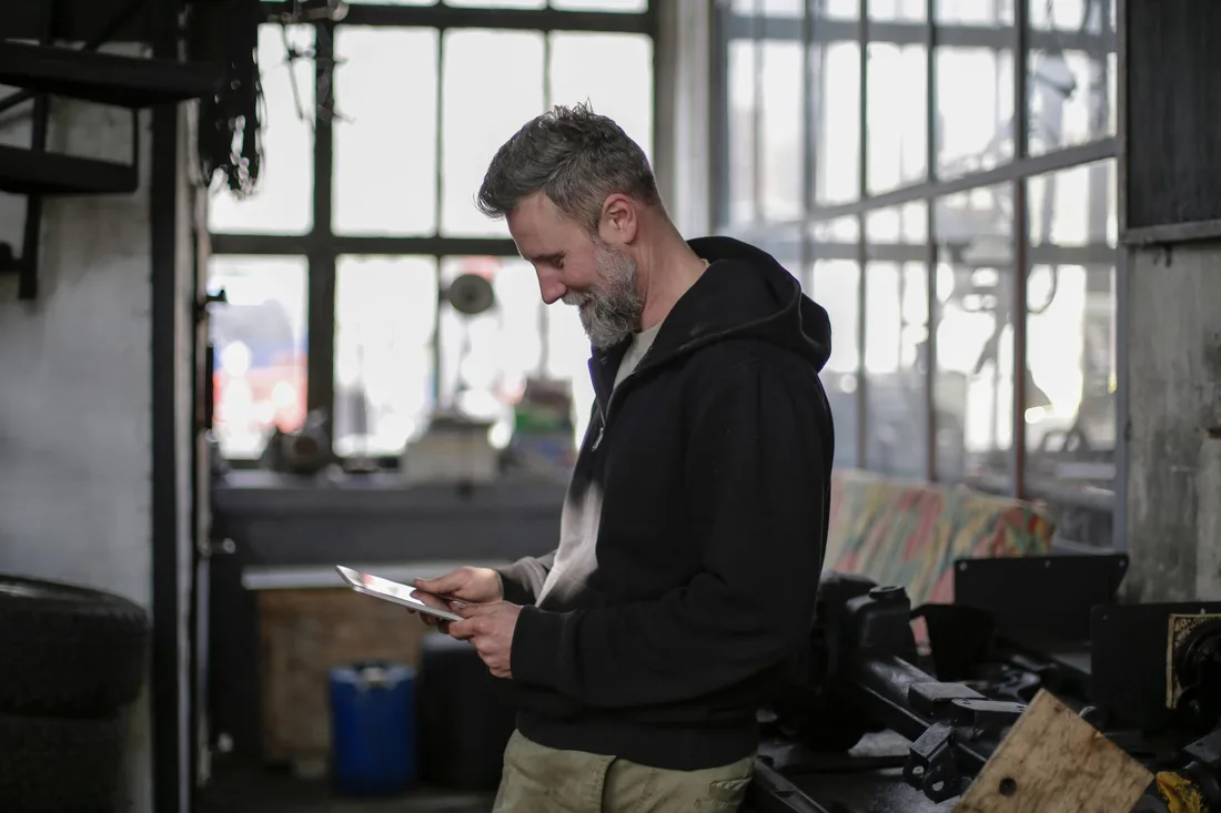 Side view of positive adult bearded male mechanic wearing black clothes standing near workbench and using tablet while working in spacious workshop. Photo by Andrea Piacquadio on Pexels