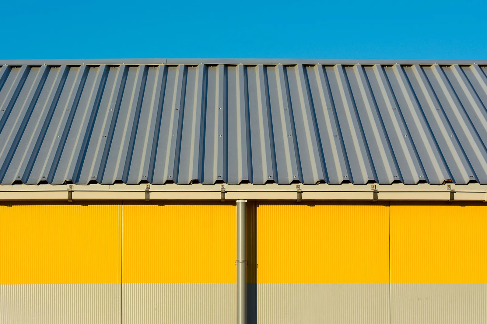 Photo of a yellow warehouse wall with a corrugated metal roof under a clear blue sky. Photo by Jan van der Wolf on Pexels