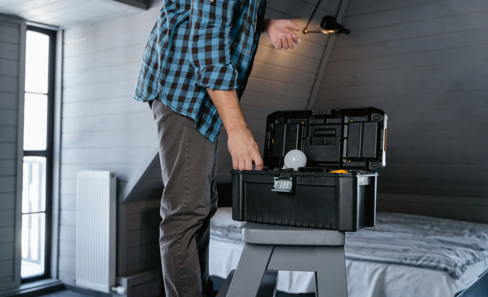 Person in plaid shirt fixing a light bulb using a toolbox in a rustic attic interior. Photo by MART  PRODUCTION on Pexels