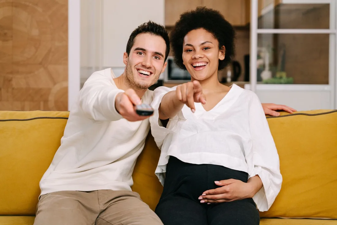 Smiling couple on a yellow sofa pointing a remote at the television. Photo by Ivan S on Pexels