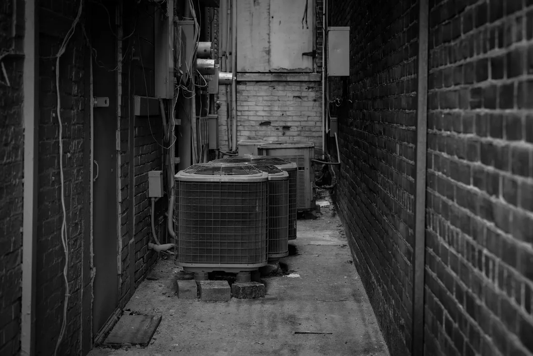 Black and white alley featuring industrial air conditioning units against a brick wall. Photo by Chris F on Pexels