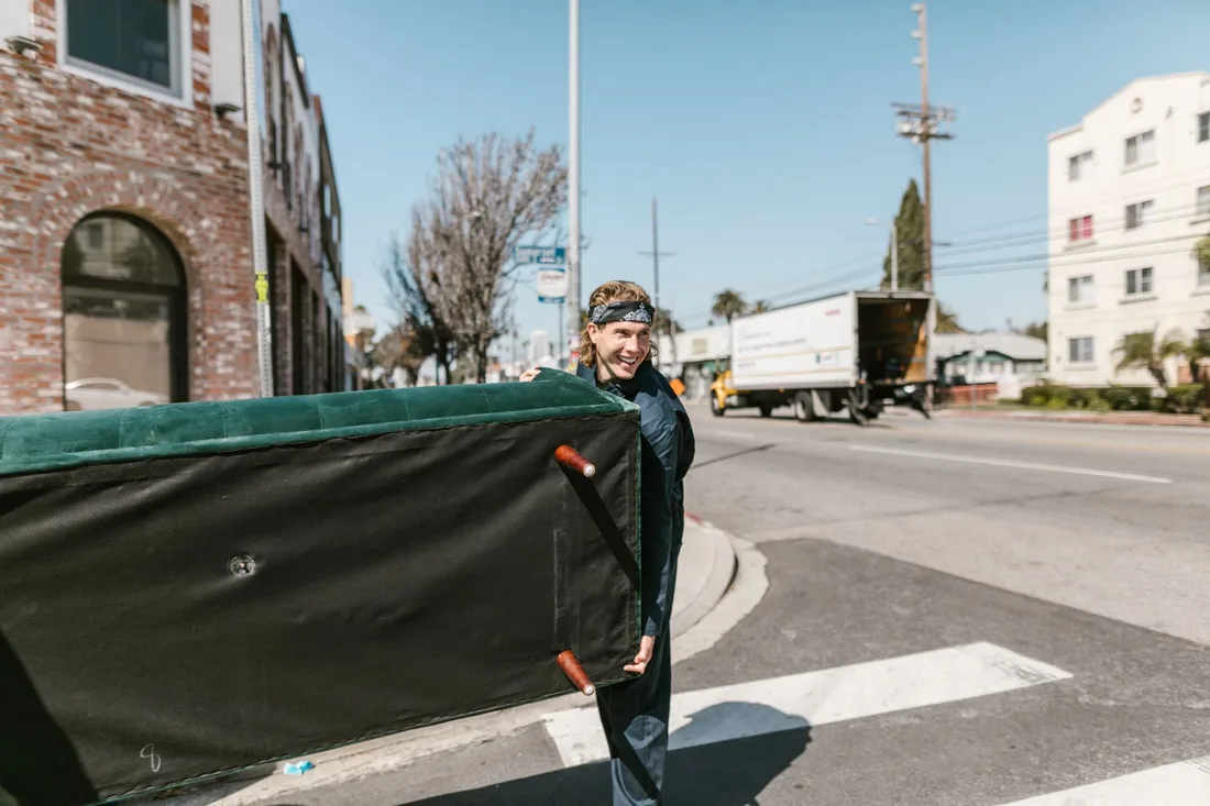 A cheerful man carries a large couch across a sunny urban street during a move. Photo by RDNE Stock project on Pexels