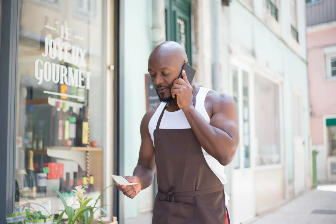 Man in apron making a business call outside a gourmet store in Portugal, checking notes. Photo by Kampus Production on Pexels