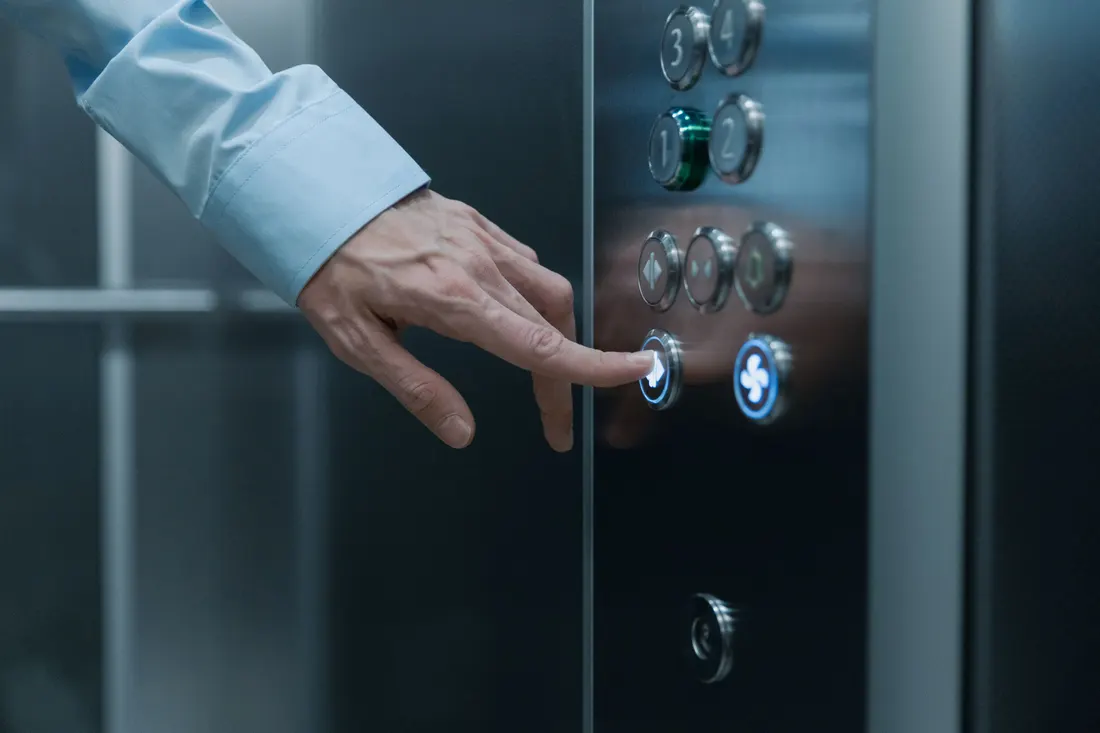 Close-up of an adult's hand in a light blue shirt sleeve pressing a lit button on a stainless steel elevator control panel inside a glass-walled car. Photo by cottonbro studio on Pexels