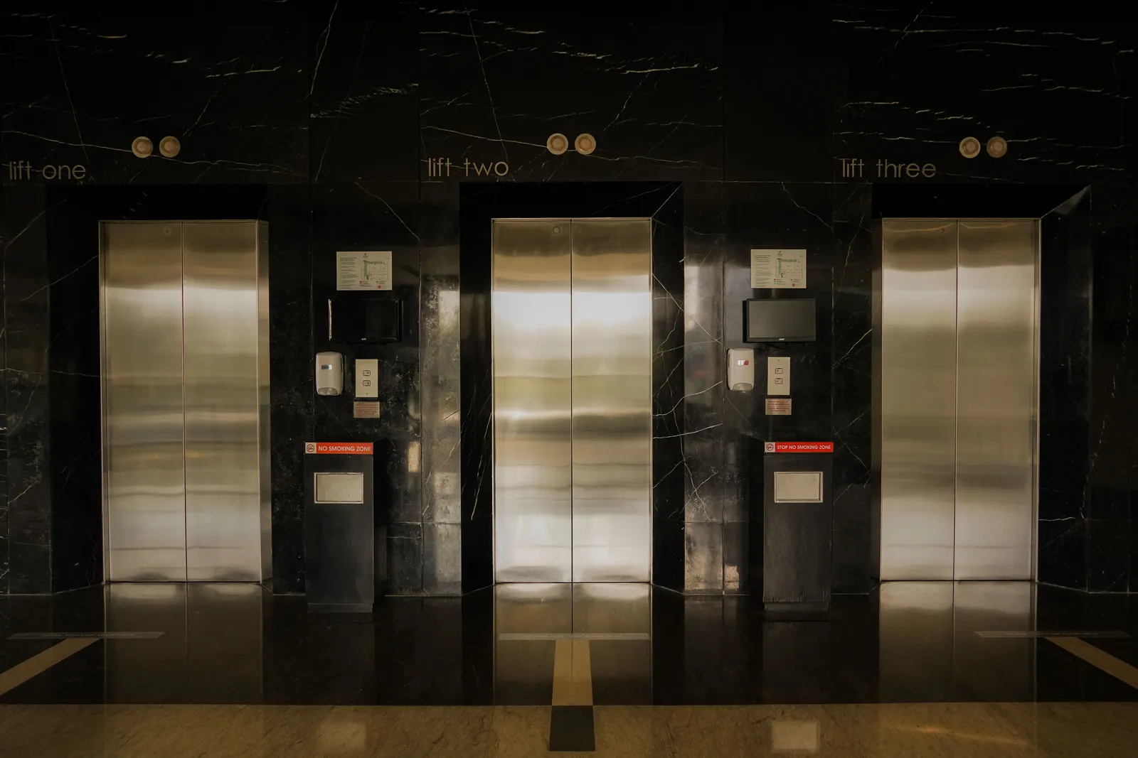 Three closed stainless steel lift doors set into a black marble lobby wall, each labelled lift one, lift two and lift three, with call buttons and small notices between them. Photo by Salman Haris on Pexels