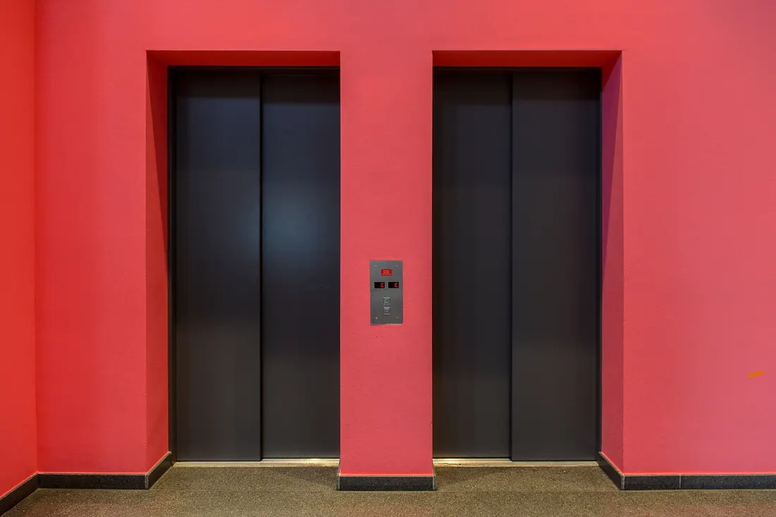Two closed dark grey elevator doors side by side in a bright red lobby wall, with a small stainless call panel between them and a speckled dark floor. Photo by Jan van der Wolf on Pexels