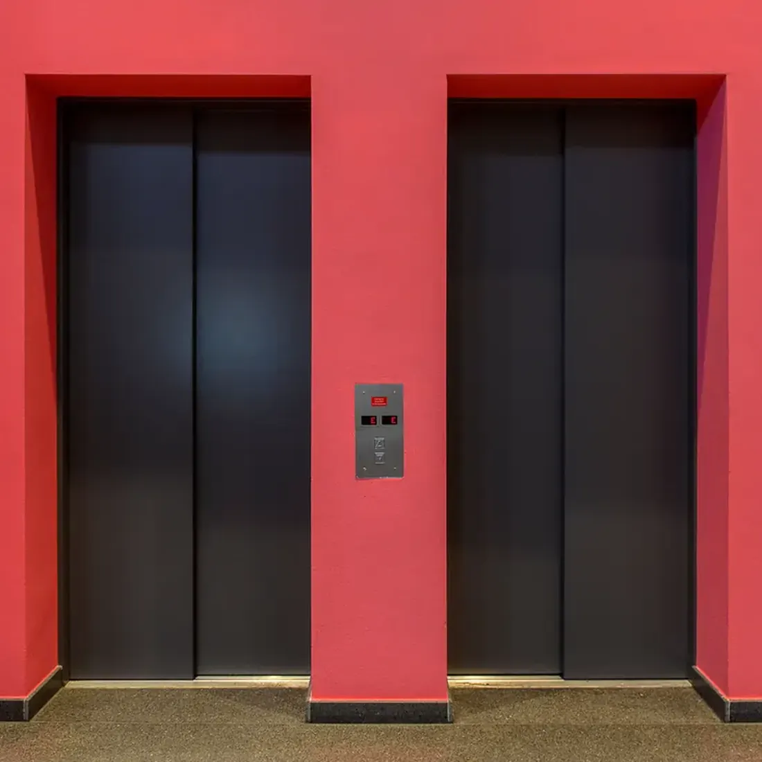 Two closed dark grey elevator doors in a bright red wall, with a small stainless call panel between them and a speckled dark floor. Photo by Jan van der Wolf on Pexels