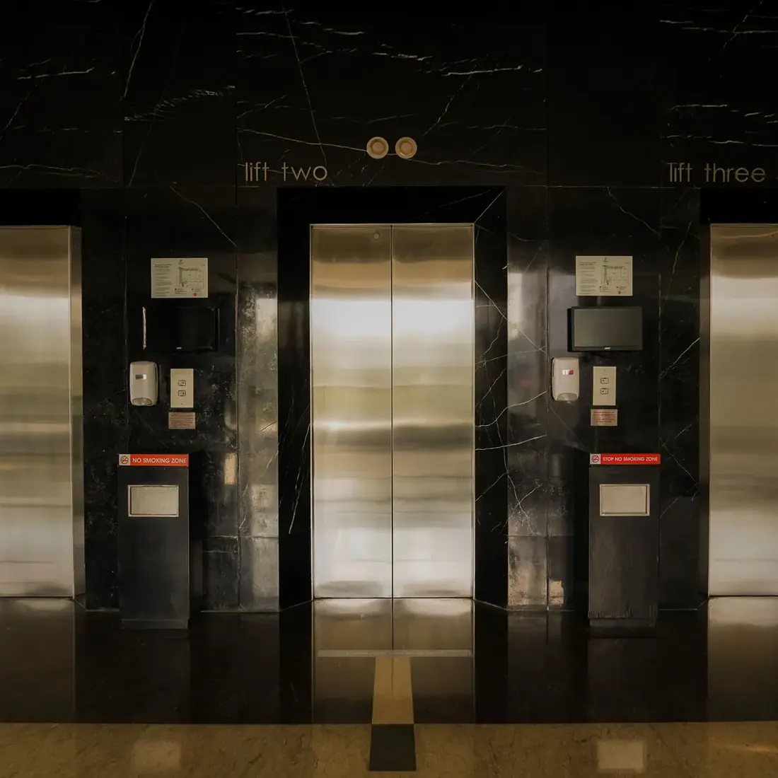 Closed stainless steel lift door labelled lift two, set in a black marble wall, with part of the neighbouring door and a no smoking notice beside it. Photo by Salman Haris on Pexels