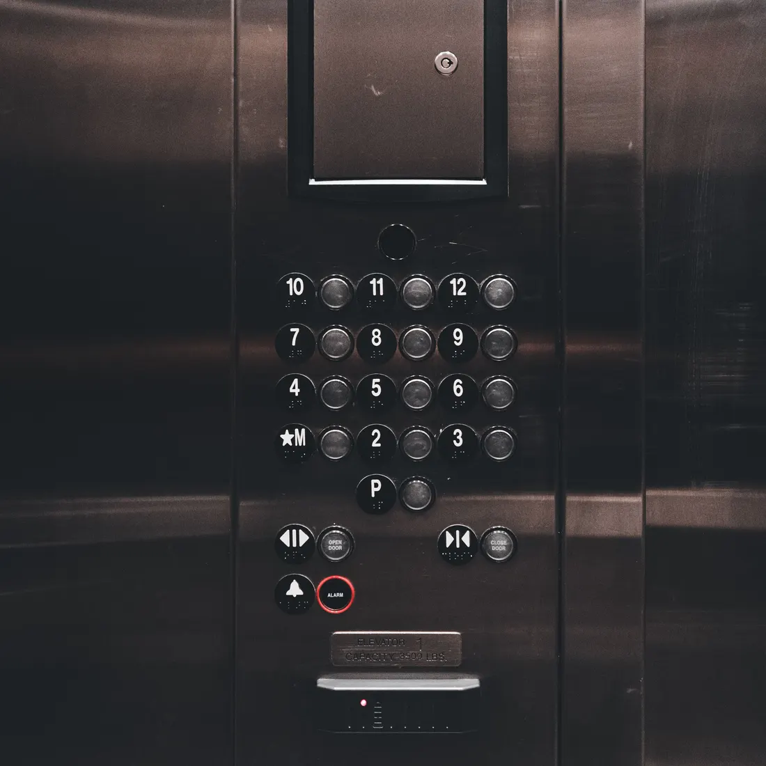 Close-up of a dark stainless steel elevator car control panel with numbered floor buttons, door open and close buttons, an alarm button and a help phone button. Photo by Erik Mclean on Pexels