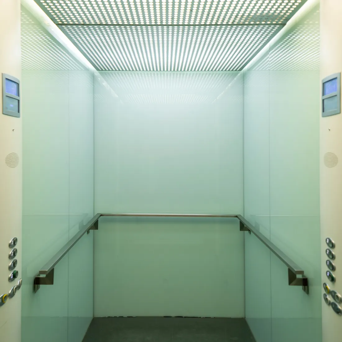 Empty pale green elevator cabin seen from the open doorway, with a stainless handrail along three walls, button columns on each side and a lit perforated ceiling. Photo by Ana Benet on Pexels