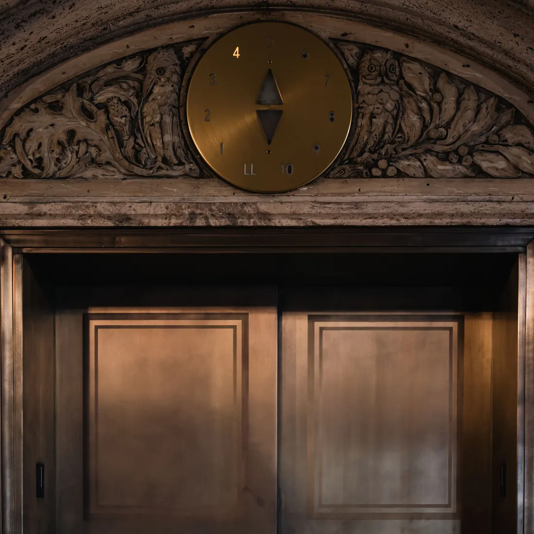 Closed bronze elevator doors beneath a carved stone arch with a round brass floor indicator dial in a historic building lobby. Photo by Chris F on Pexels