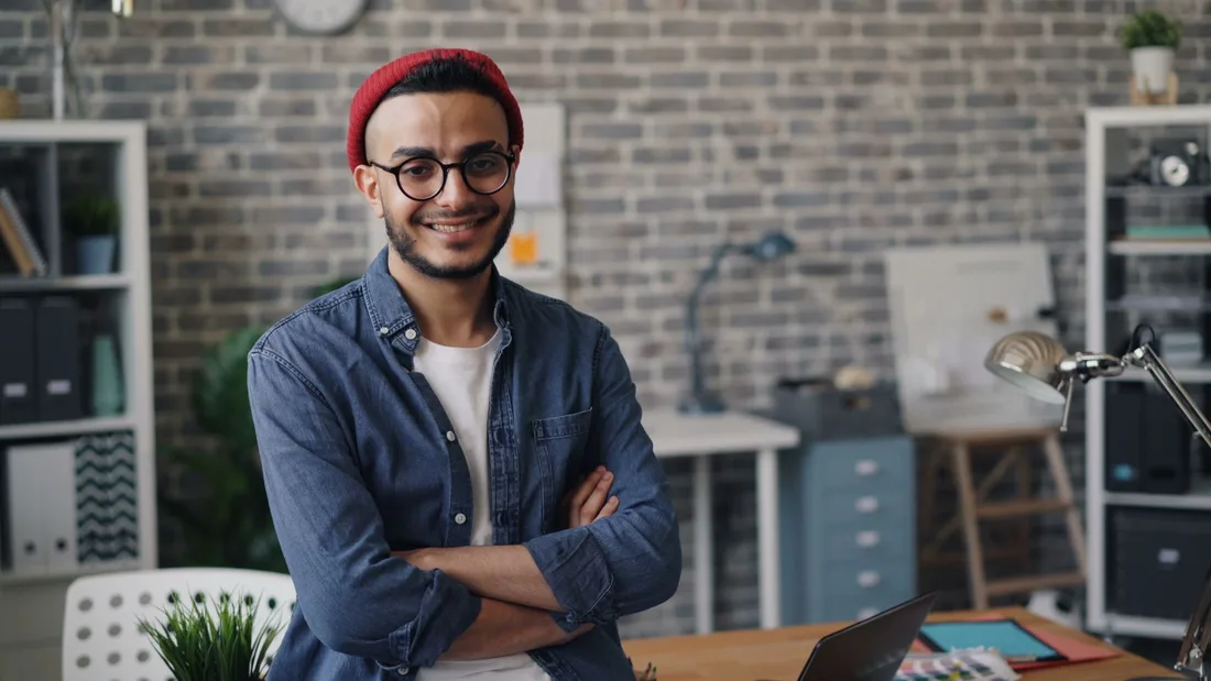 Smiling young man standing confidently in a modern office with a brick wall background. Photo by Vitaly Gariev on Pexels