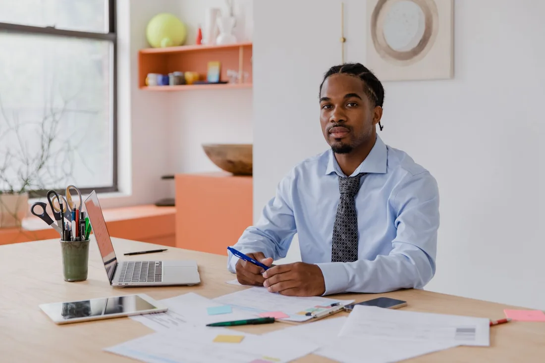 A professional businessman with a beard and braided hair sits at his desk with a laptop and documents, working diligently. Photo by RDNE Stock project on Pexels