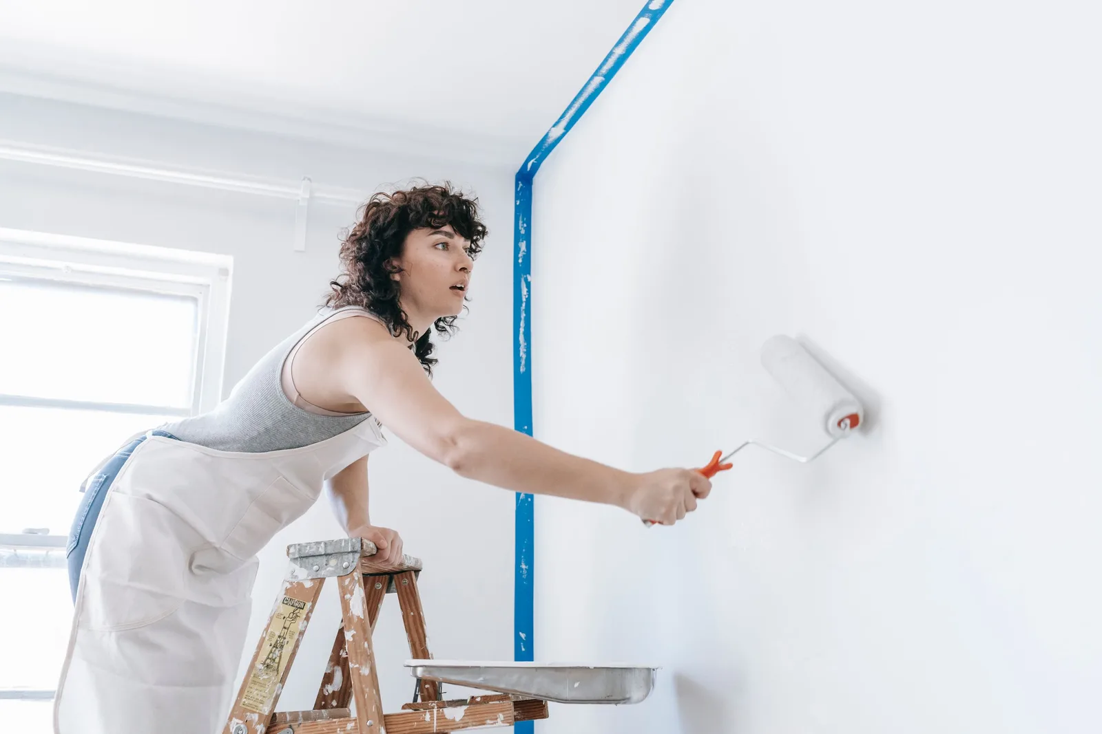 Painter in an apron rolling white paint onto a wall beside a ladder and blue tape. Photo by Blue Bird on Pexels