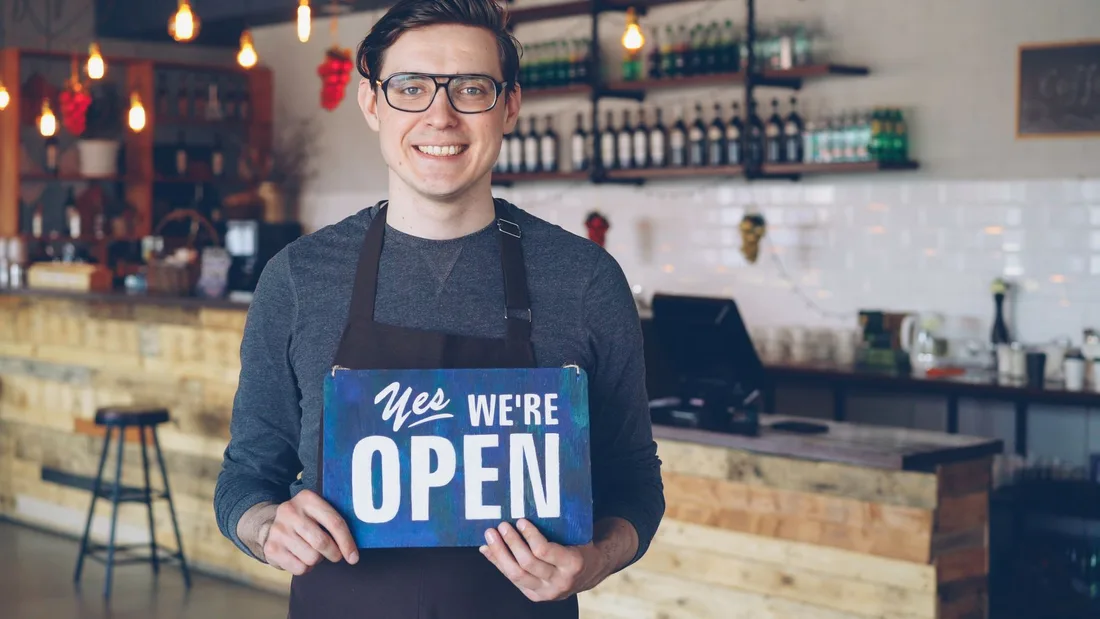 Friendly barista welcomes customers holding an open sign in a cozy cafe setting. Photo by Vitaly Gariev on Pexels