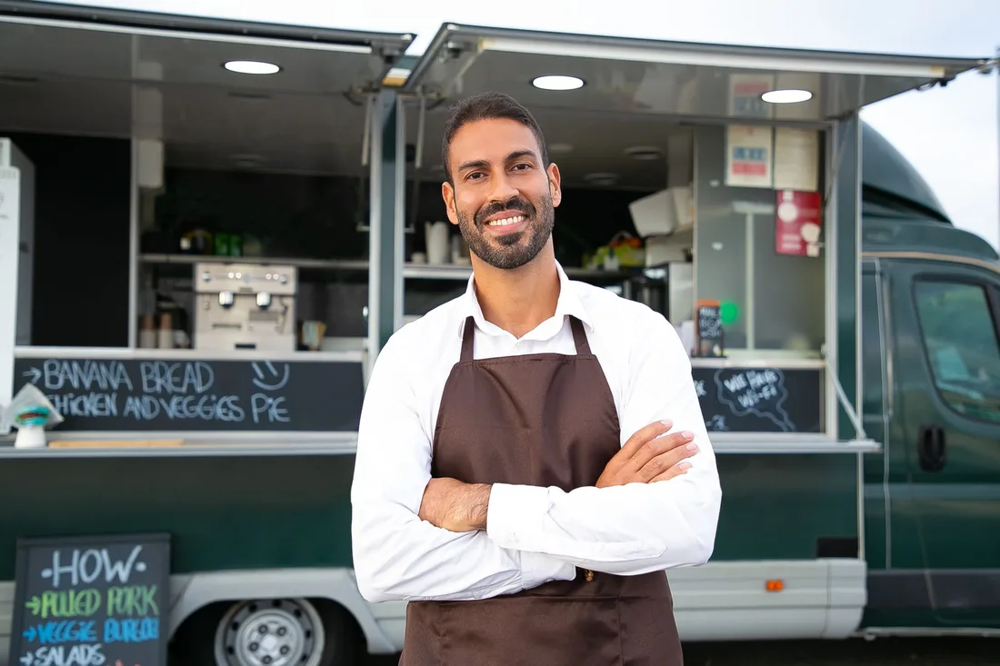 Smiling young ethnic male waiter in apron standing with arms folded near modern food truck and looking at camera contentedly. Photo by Kampus Production on Pexels