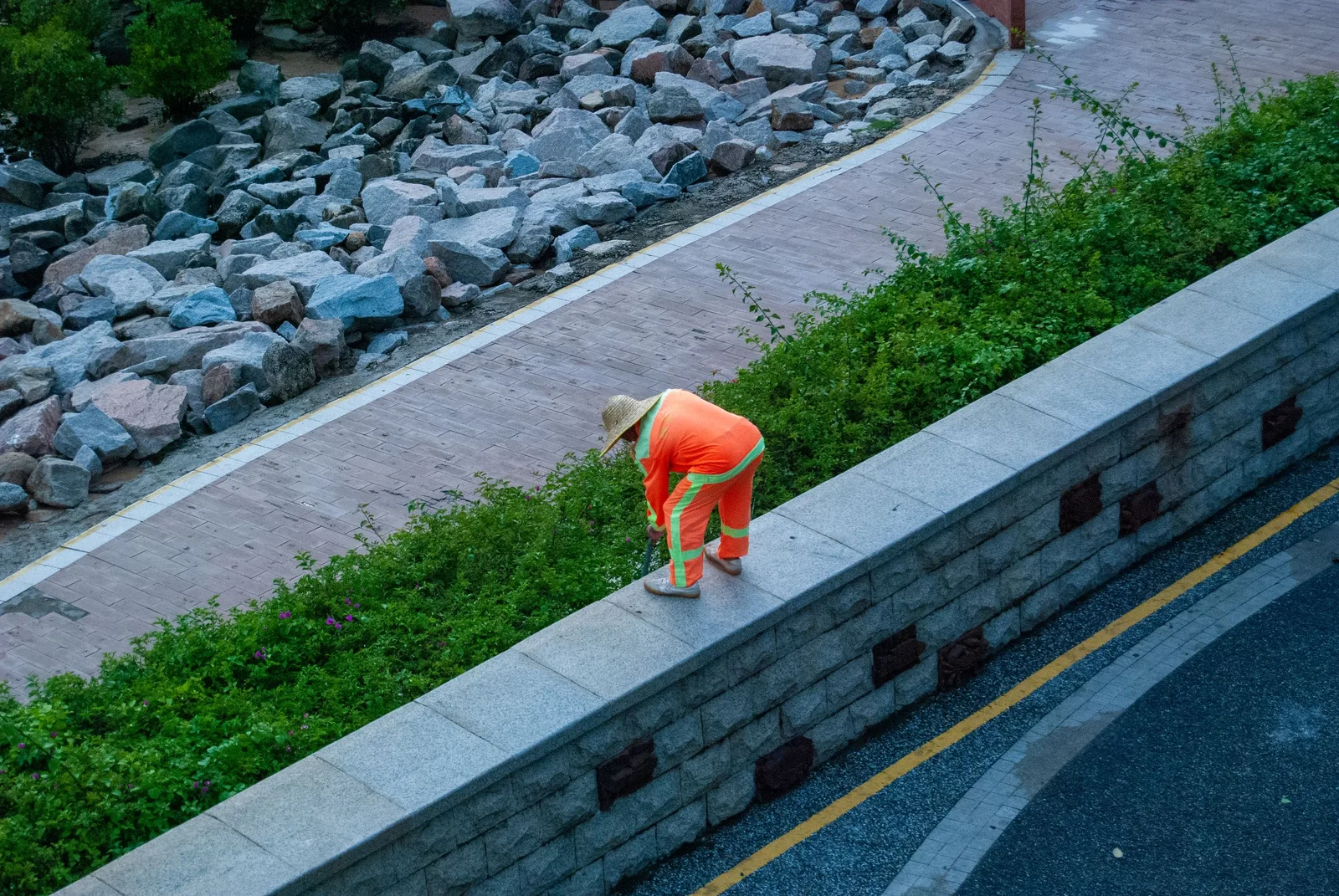 City gardener in orange uniform tending to plants on a stone wall in a park. Photo by Da Na on Pexels