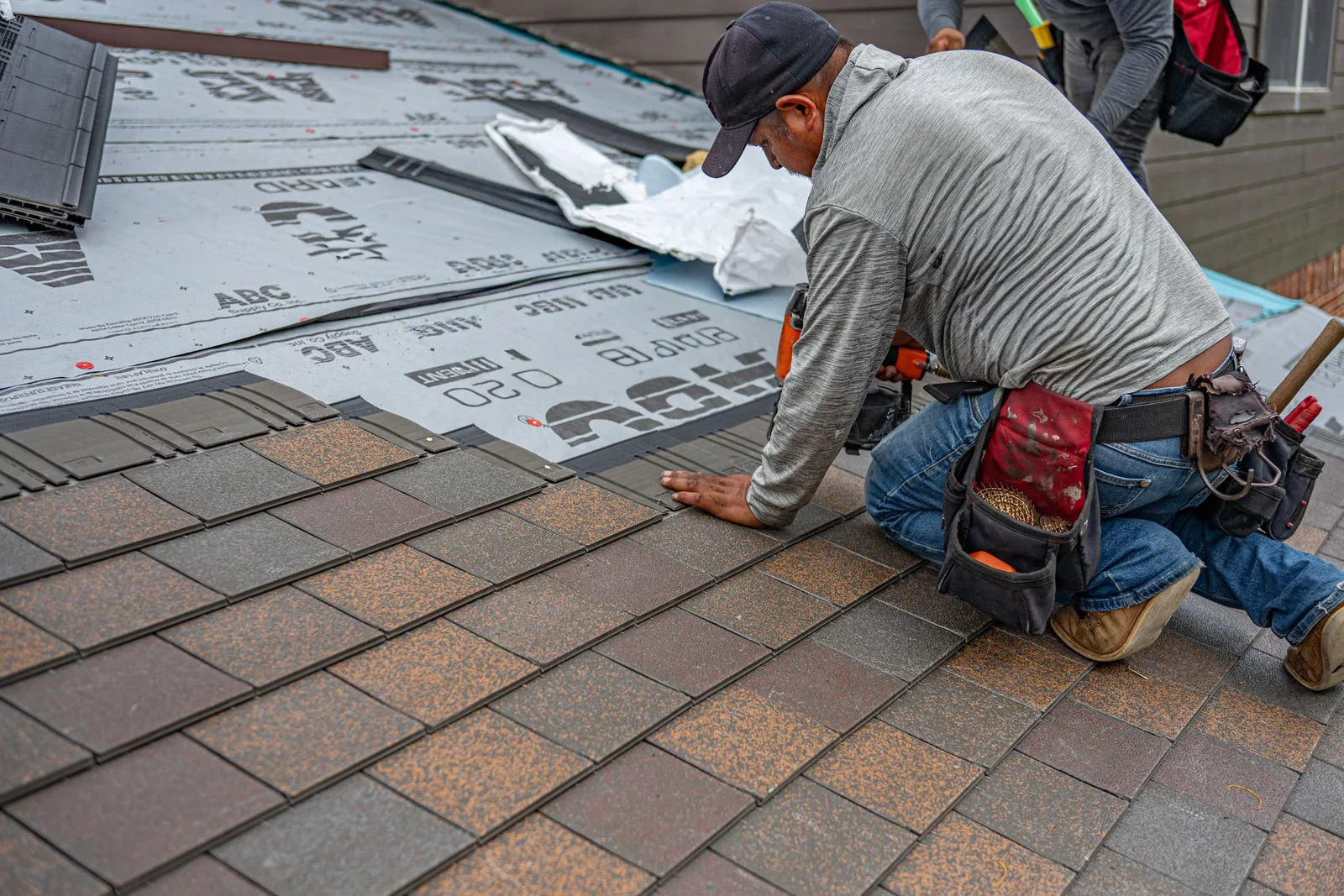 Roofer kneeling on a roof installing asphalt shingles over underlayment with a tool belt. Photo by Ryan  Stephens on Pexels