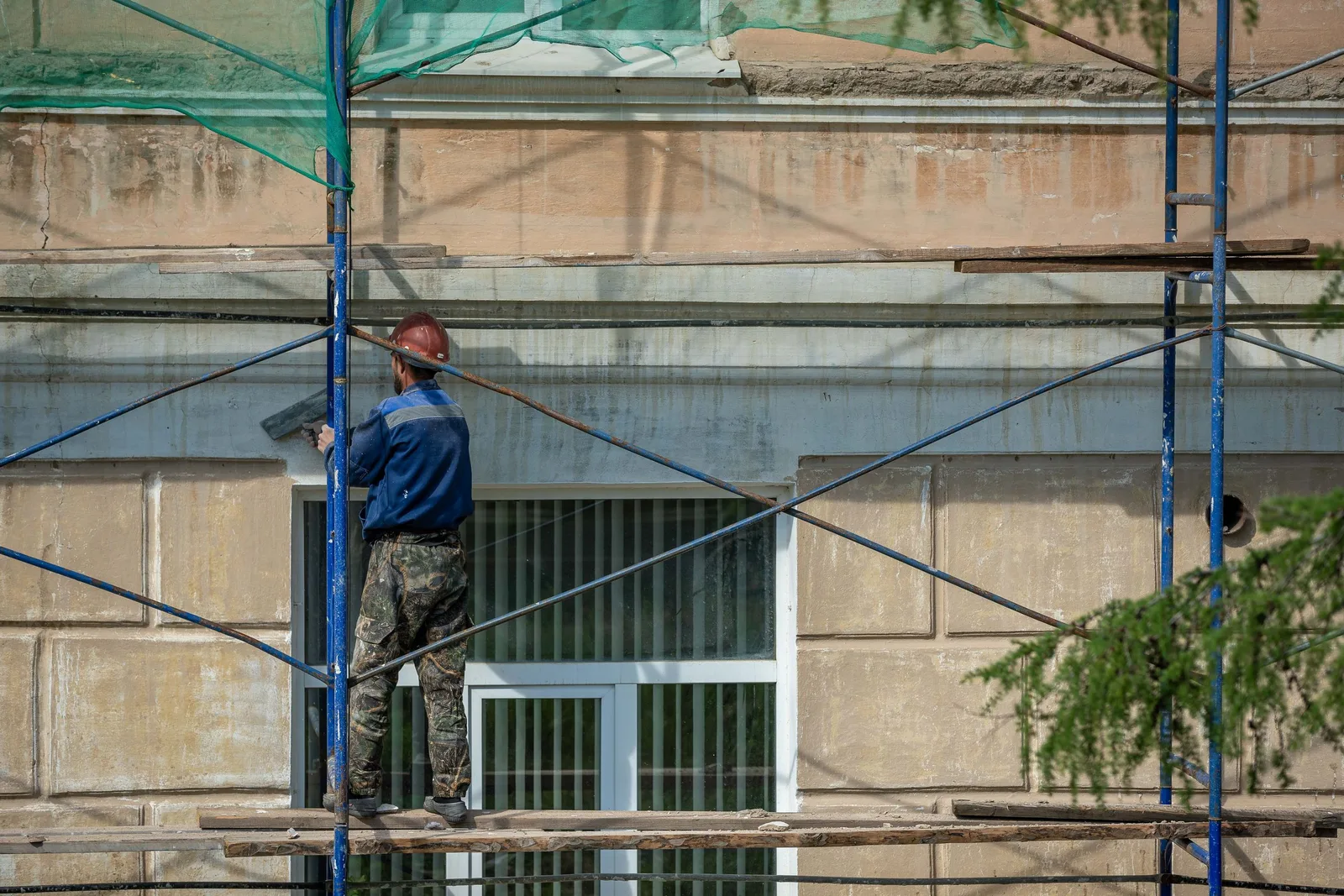 A construction worker in a hard hat repairs a building facade on scaffolding in an urban setting. Photo by Sergei Starostin on Pexels